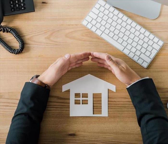 A cut out of a paper home, with arms circled over the home on a desk.